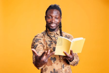 Photo for African american person being focused on fictional tale for leisure activity, enjoying reading literature book in studio over yellow background. Positive man studying for university homework - Royalty Free Image