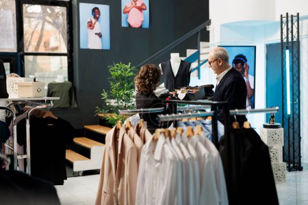 Téléchargez les photos : Un employé du showroom aide un homme âgé à choisir la bonne cravate pour une tenue formelle dans une boutique moderne, discutant du tissu des accessoires. Client âgé achetant des marchandises à la mode dans un magasin de vêtements - en image libre de droit
