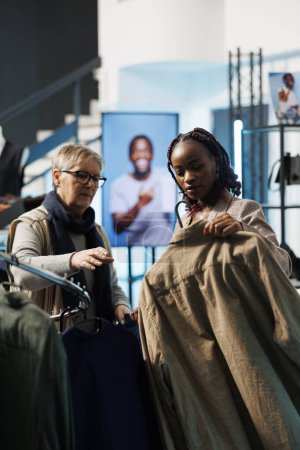 Photo for African american employee helping elderly client choosing brown shirt for husband in modern boutique. Senior woman shopping for casual wear, buying fashionable merchandise in clothing store - Royalty Free Image