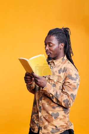 Photo for Concentrated clever person holding literature book, reading fiction story in studio over yellow background. Intellectual african american man studying before start working at university homework - Royalty Free Image