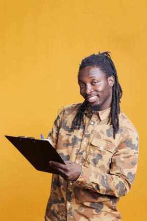 Photo for Smiling man taking notes on notebook files using pen in studio. African american person writing on clipboard papers, signing documents about business project, analyzing job checklist - Royalty Free Image