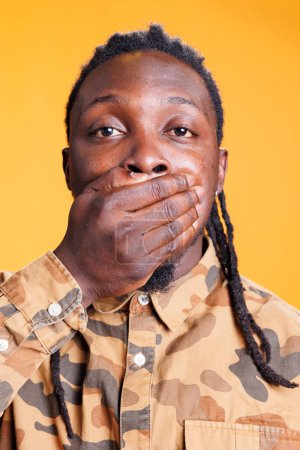 African american man covering mouth with palms, doing doing three wise monkeys symbolic gesture in studio over yellow background. Young adult showing silence gesture, not speaking concept