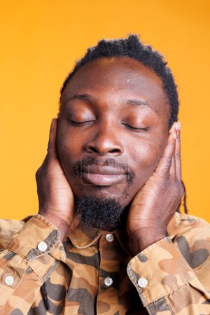 Photo for African american man covering ears with palms, doing three wise monkeys gesture in front of camera over yellow background. Person with serious expression not listening to noise and not speaking - Royalty Free Image