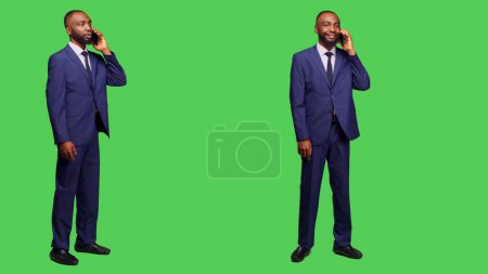 Photo for Young businessman in suit talking on phone call, standing over full body greenscreen. Male office employee having remote conversation using smartphone line in studio, enjoying telephone chat. - Royalty Free Image