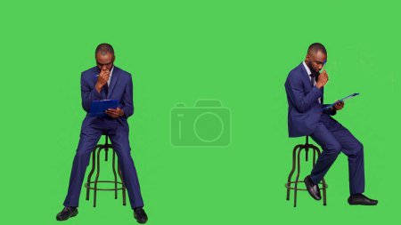 Téléchargez les photos : Un Afro-Américain réfléchissant à des notes d'affaires sur des papiers, réfléchissant à de nouvelles idées sur des dossiers de presse-papiers. Jeune homme d'affaires assis sur une chaise en studio, se sentant réfléchi et coûteux. - en image libre de droit