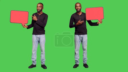 Photo for Male model holding red blank speech bubble on camera, creating presentation with empty copyspace cardboard icon. Young adult showing mockup billboard over green screen backdrop. - Royalty Free Image