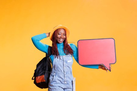 Photo for Joyful woman holding speech bubble in studio, showing cardboard with isolated copyspace on camera. Young adult creating advertisement with empty red billboard, urban holiday trip. - Royalty Free Image