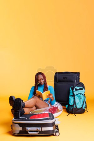 Téléchargez les photos : Livre de lecture touristique afro-américain, assis sur le sol avec gonflable et bagages en studio. Jeune femme profitant d'une conférence pour une activité de loisirs, voyageant à l'étranger sur citybreak. - en image libre de droit