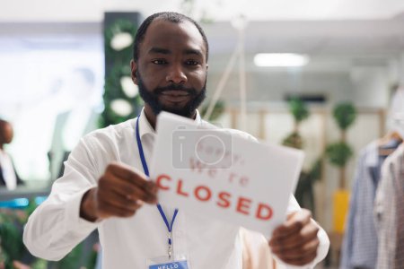 Photo for Fashion garment department store african american employee hanging closed sign on front door. Apparel shop worker holding signboard informing customers about work day end - Royalty Free Image