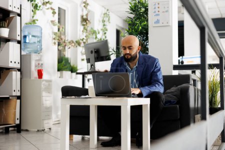 Téléchargez les photos : Homme d'affaires arabe travaillant sur ordinateur portable dans le bureau de démarrage d'entreprise. Homme gestionnaire professionnel de planification de projet stratégie de marketing de vente tout en étant assis sur le canapé dans l'espace de coworking moderne - en image libre de droit
