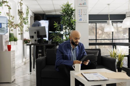 Téléchargez les photos : Homme d'affaires arabe analysant rapport financier sur la table numérique tout en étant assis sur le canapé dans le bureau de démarrage d'entreprise. Employé de l'entreprise travaillant au marketing des médias sociaux dans un espace de coworking moderne - en image libre de droit