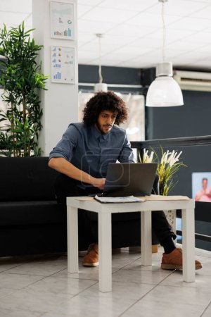 Téléchargez les photos : Jeune homme arabe analysant les statistiques financières sur ordinateur portable tout en travaillant dans le démarrage de petites entreprises bureau à aire ouverte. Stratégie de projet de l'entreprise de planification d'homme d'affaires concentré - en image libre de droit