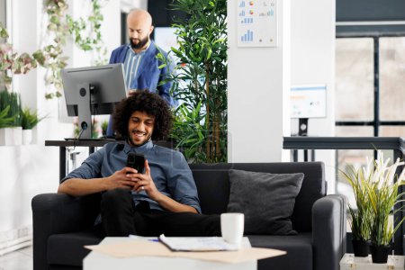Téléchargez les photos : Joyeux entrepreneur arabe bavarder en ligne sur smartphone tout en étant assis sur le canapé dans le bureau de démarrage d'entreprise. Rire homme d'affaires en regardant une vidéo drôle sur téléphone mobile dans l'espace de coworking - en image libre de droit