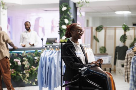 Photo for Smiling african american clothes store assistant waiting for customers while standing near apparel rack. Cheerful young fashion boutique shop professional employee working - Royalty Free Image