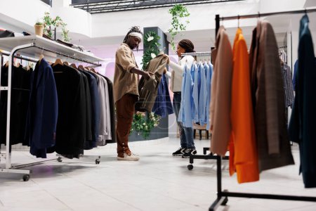 Téléchargez les photos : Afro-américain homme et femme acheter des vêtements dans le centre commercial et de décider entre deux vestes sur cintres. Petit ami et petite amie clients choisissant des vêtements formels en magasin - en image libre de droit