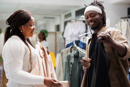 Téléchargez les photos : Couple afro-américain choisissant veste formelle pour une occasion spéciale dans un magasin de vêtements. Souriant jeune homme montrant blazer moderne à la femme et demandant à essayer dans le centre commercial - en image libre de droit