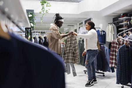 Foto de Cliente afroamericano recibiendo ayuda de su novia en la elección de la camisa formal en la boutique de moda. Pareja de compras en la tienda de ropa y comparar ropa masculina en perchas - Imagen libre de derechos