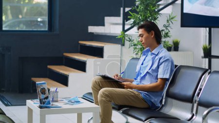 Photo for Asian patient sitting on chiar in hospital waiting area, filling medical documents with nurse before attenting examination. Young adult signing insurance form during checkup visit. Medicine service - Royalty Free Image