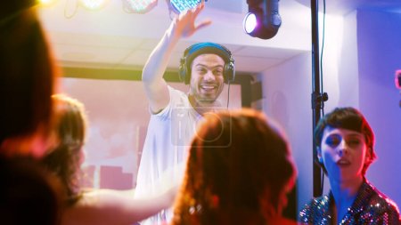 Photo for Cool DJ clubbing with people on stage, mixing electronic sounds on audio station panel to create party atmosphere at club. Young adult partying and jumping on dance floor, entertainment. - Royalty Free Image