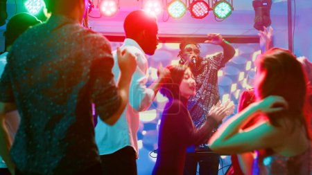 Diverse group of friends at nightclub, dancing on electronic mix from DJ music station in discotheque. Funky people partying and enjoying modern audio sounds, colorful discolights.