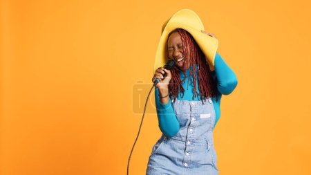 Foto de Joven mujer feliz cantando en el micrófono, haciendo karaoke delante de la cámara. Alegre cantante talentoso que tiene actuación musical en vivo en el estudio, de pie sobre fondo naranja. - Imagen libre de derechos