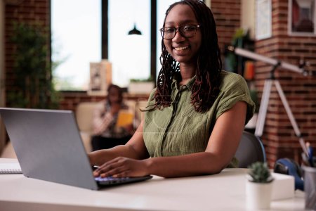 Photo for Portrait of african american freelancer smiling at camera while typing on laptop computer in home living room. Casual entrepreneur working remote posing happy while roommate relaxes on sofa. - Royalty Free Image