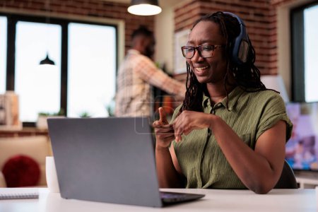 Téléchargez les photos : Femme souriante avec écouteurs sans fil regarder spectacle de divertissement sur ordinateur portable tout en étant assis au bureau. pigiste afro-américain regardant contenu drôle de médias sociaux sur ordinateur portable. - en image libre de droit