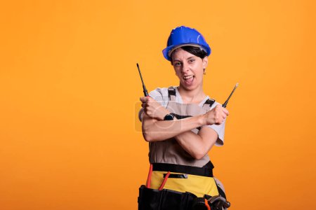 Photo for Energetic female construction worker looking at camera holding screwdrivers in studio. Happy woman builder wearing overalls and protective helmet with screwing tools, yellow background. - Royalty Free Image