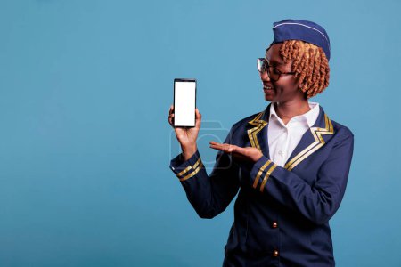 Photo for Female flight attendant wearing uniform holds cell phone with empty screen for advertising isolated on blue background. Professional african american stewardess looking at camera in studio shot. - Royalty Free Image