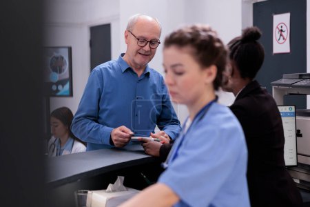 Photo for Senior patient standing at registration counter paying with credit card for medication treatment and consultation using contactless payment in hospital waiting room. Health care service and support - Royalty Free Image