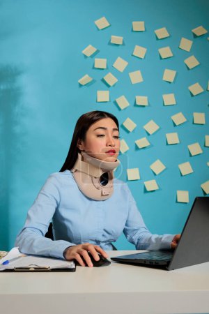 Photo for Asian woman with cervical collar on neck using laptop computer at modern office desk. Female employee with injury making an effort to finish sales reports in startup company. - Royalty Free Image