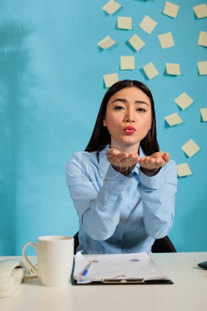 Photo for Asian professional woman showing positive feelings at work place, throwing kisses in the air while working on report. Affectionate female employee showing affectionate emotions in modern office. - Royalty Free Image