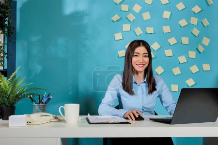Photo for Asian lady planning report on laptop computer before monthly statistics meeting. Smiling businesswoman working from desk in modern office with adhesive papers attached to the wall. - Royalty Free Image