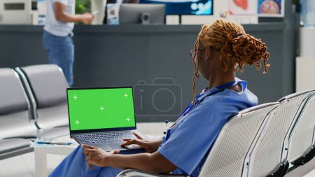 Téléchargez les photos : Infirmière tenant un ordinateur portable avec écran vert dans la salle d'attente, en utilisant un modèle de maquette isolé à la réception de l'hôpital. Jeune femme travaillant avec clé chromatique vierge et copyspace. Trépied. - en image libre de droit