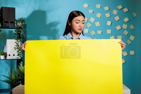 Téléchargez les photos : Femme asiatique optimiste tenant signe en carton jaune tout en posant dans un bureau moderne sur fond bleu. Employée confiante montrant affiche publicitaire avec espace de copie. - en image libre de droit