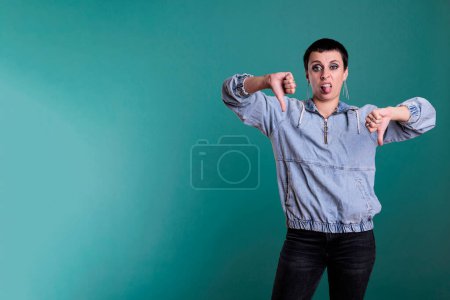 Photo for Negative angry woman doing thumbs down gesture while posing in studio over isolated background. Dissatisfied female giving dislike sign and feeling disappointed, gesturing negativity symbol. - Royalty Free Image