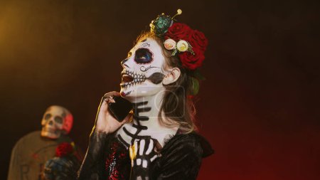 Photo for Lady of dead talking on phone call with smartphone, wearing flowers crown and cavalera catrina halloween costume. Woman dressed as santa muerte to celebrate mexican culture holiday. Handheld shot. - Royalty Free Image