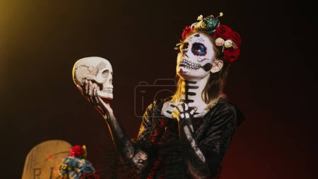 Foto de La cavalera catrina hablando con cráneo en estudio, actuando de miedo y horror para celebrar la tradición mexicana de halloween. Santa muerte vestida con espeluznante disfraz de horror con arte corporal. Disparo de mano. - Imagen libre de derechos