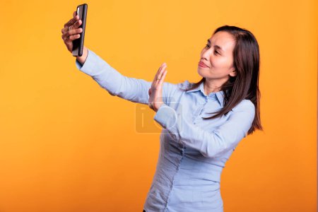 Foto de Mujer atractiva asiática saludando a personas remotas durante la reunión de videocall de teléfono móvil, disfrutando de la conversación en el estudio sobre fondo amarillo. Adulto seguro divirtiéndose con un amigo durante la llamada - Imagen libre de derechos