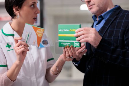 Photo for Pharmacy assistant recommending vitamin and aspirin to client, helping to choose anti inflammatory pills in drugstore. Apothecary elderly customer talking with pharmacist about painkiller prescription - Royalty Free Image