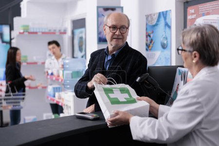 Photo for Senior man taking medications order from pharmacist at checkout. Elderly client buying supplements and disease remedy in pharmacy store, consulting with drugstore employee at cash register - Royalty Free Image