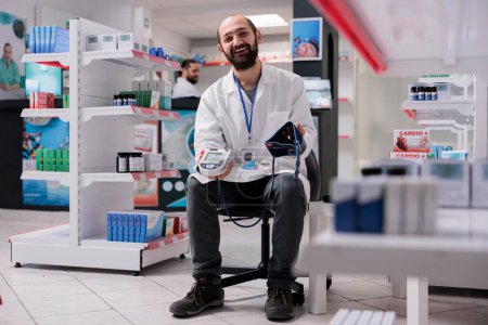 Photo for Smiling pharmacist holding medical tonometer ready to measure customers hypertension during checkup visit in pharmacy. Drugstore filled with supplements and drugs, medicine support service - Royalty Free Image