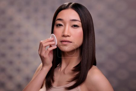 Téléchargez les photos : Femme asiatique en utilisant du coton pour enlever la crème de fond de teint visage, nettoyer le visage et regarder la caméra. Belle jeune modèle montrant la procédure quotidienne de routine de soins de la peau pour une peau saine - en image libre de droit
