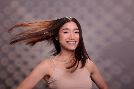 Téléchargez les photos : Heureuse femme asiatique avec des cheveux flottants brillants après les procédures de routine de soins capillaires souriant et regardant la caméra. Confiant optimiste jeune femme avec portrait coiffure lisse - en image libre de droit