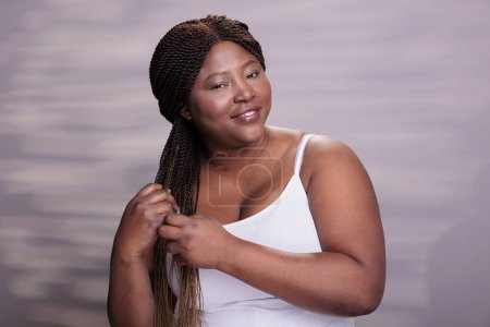 Foto de Mujer afroamericana hermosa natural haciendo peinado trenzas, vistiendo top blanco, sonriendo y mirando a la cámara. Atractivo modelo positivo corporal que muestra el cuidado del cabello procedimiento diario - Imagen libre de derechos