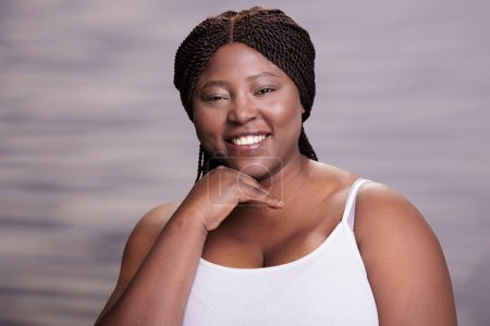 Photo for Attractive smiling african american curvy young woman holding hand on face portrait. Cheerful body positive lady putting arm on cheek, looking at camera and posing in studio - Royalty Free Image