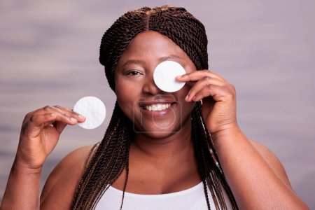 Foto de Mujer afroamericana quitándose los ojos maquillarse con almohadillas de disco de algodón, haciendo rutina de cuidado de la piel para una piel sana. Sonriente más tamaño modelo cara de limpieza de cosméticos decorativos retrato - Imagen libre de derechos