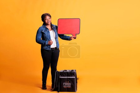 Photo for African american pizzeria courier holding red speech bubble advertisting takeout food service while standing in studio with yellow background. Restaurant courier carrying takeaway thermal backpack - Royalty Free Image