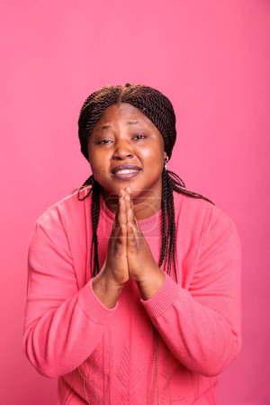 Téléchargez les photos : Portrait d'une femme religieuse afro-américaine priant Dieu de lui demander pardon tout en restant debout dans un studio avec un fond rose. Jeune adulte tenant les paumes ensemble dans la prière. Concept de religion - en image libre de droit