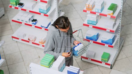Téléchargez les photos : Asian male client looking at boxes of pills in pharmacy shop, reading package to buy prescription medicine. Checking medicaments and supplements before taking treatment from shelves. Handheld shot. - en image libre de droit